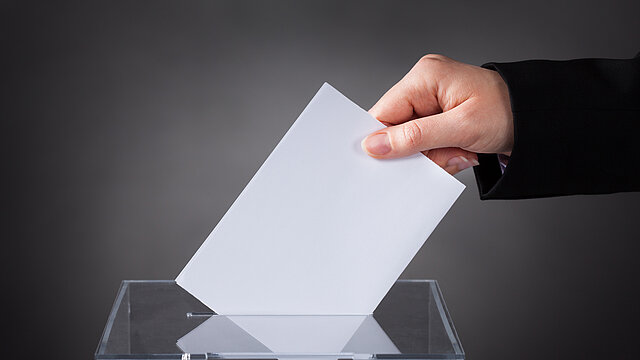 Zdjęcie dłoni wrzucającej białą kartkę do przezroczystej urny.